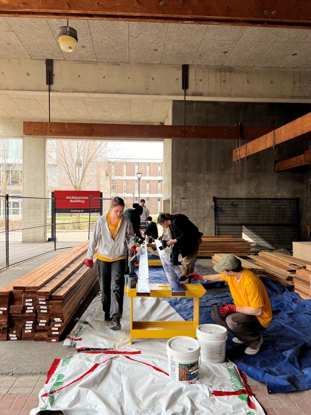 Students staining long wood pieces for the HUB structure under the architecture building overhang.