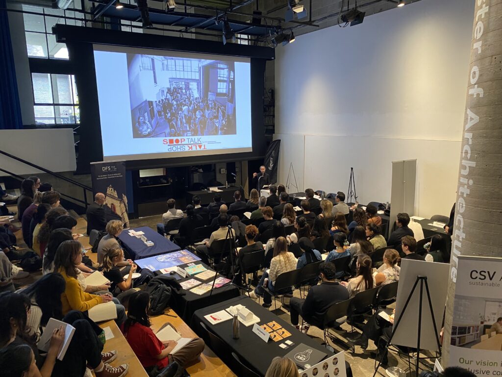 Wide view of a packed auditorium during a Shop Talk lecture, with posters and booths visible around the Pit.