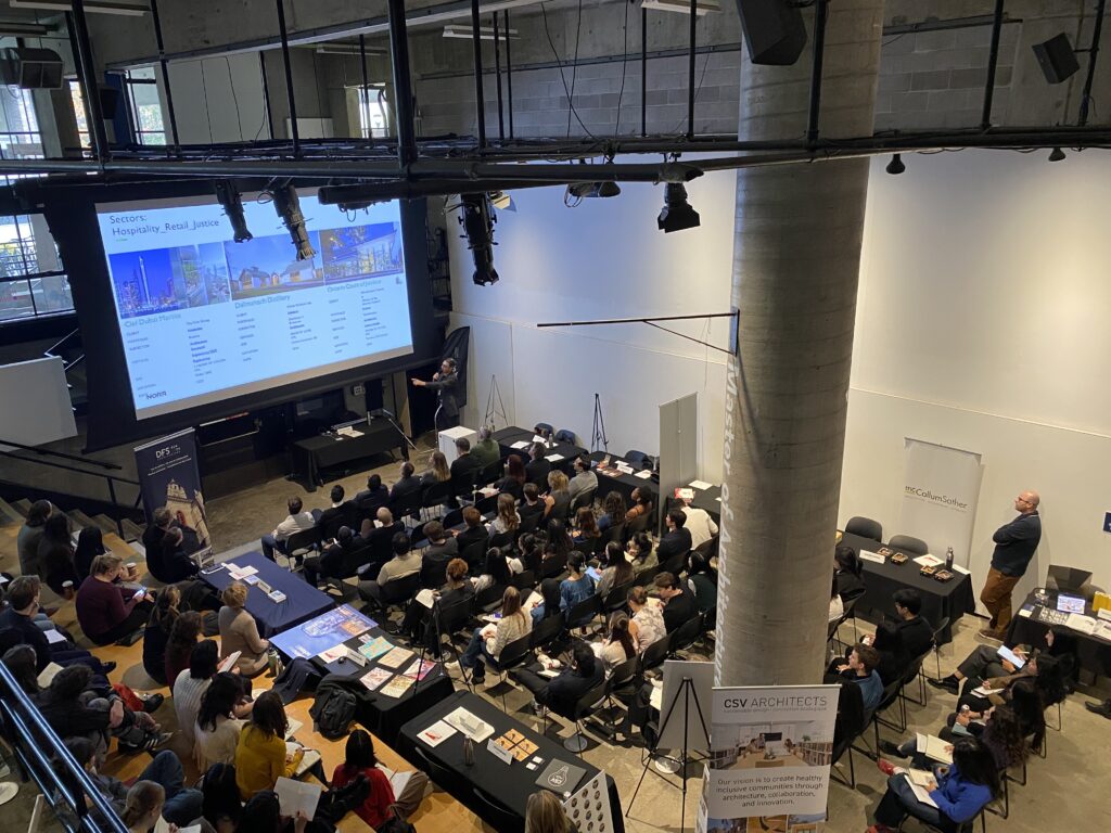 Speaker presenting architectural projects on large screen to seated audience at the Pit