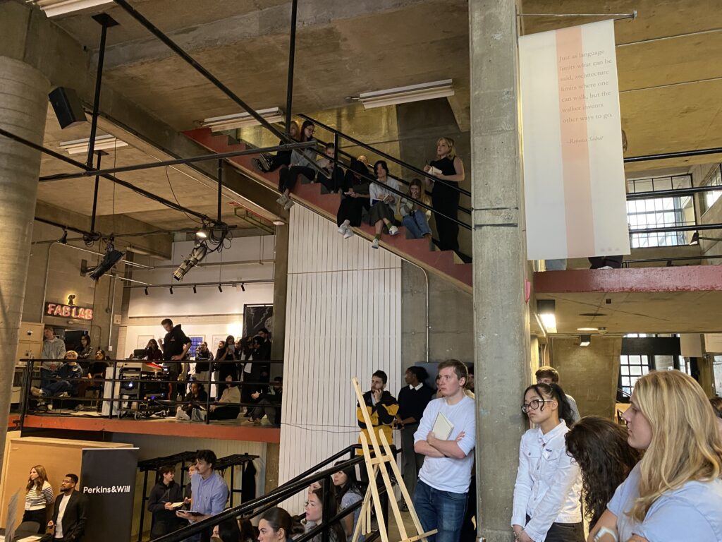 Architecture students watching a speaker presenting, with peers seated on stairs and standing along the upper railings.