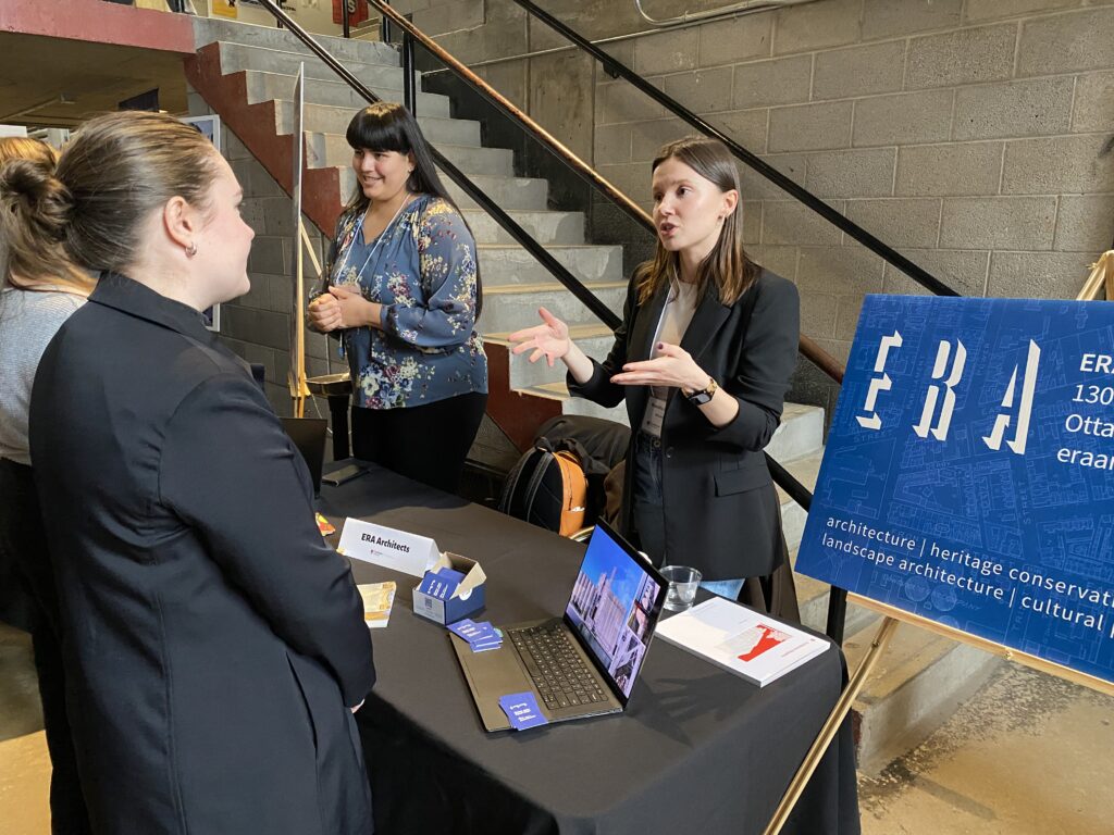 Two ERA Architects representatives speak with students at a career fair table near a staircase and poster.
