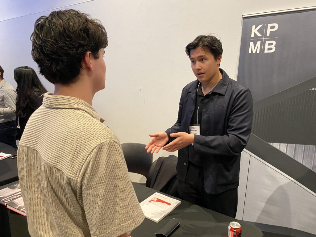 A representative from KPMB speaks with a student, gesturing while standing beside a large banner with the firm's logo.