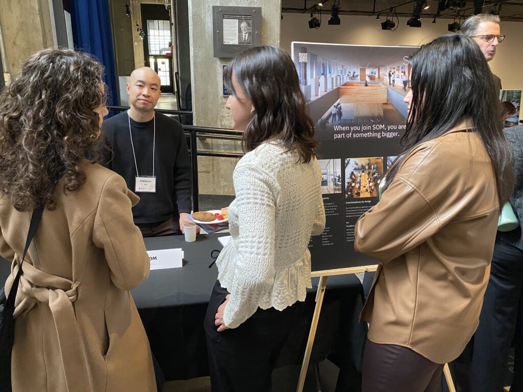 An SOM representative stands behind a booth talking with students in front of a large firm display board.