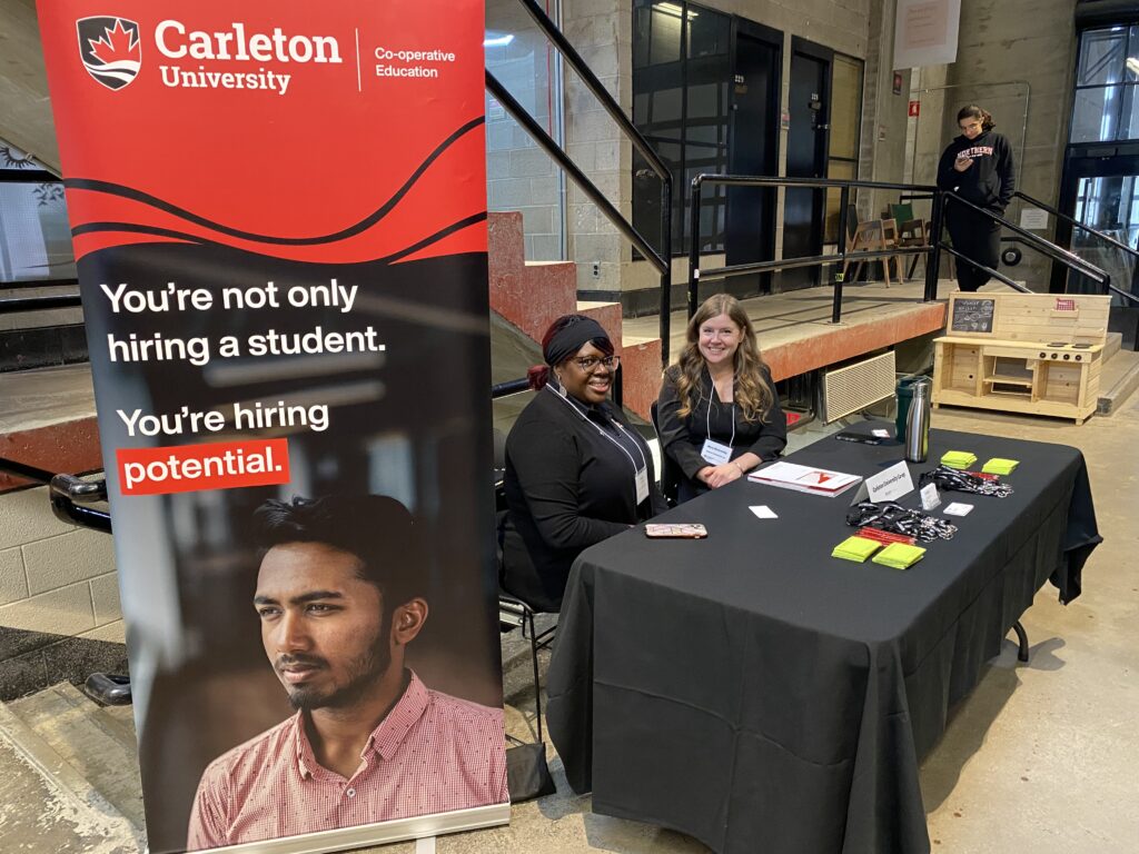 Two women sit behind a Carleton Co-op Education booth with a red banner reading “You’re hiring potential.”