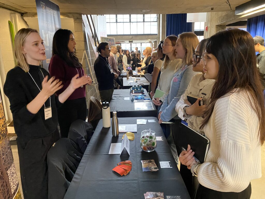 Two representatives at a career fair booth talk to a group of students holding notebooks and listening attentively.