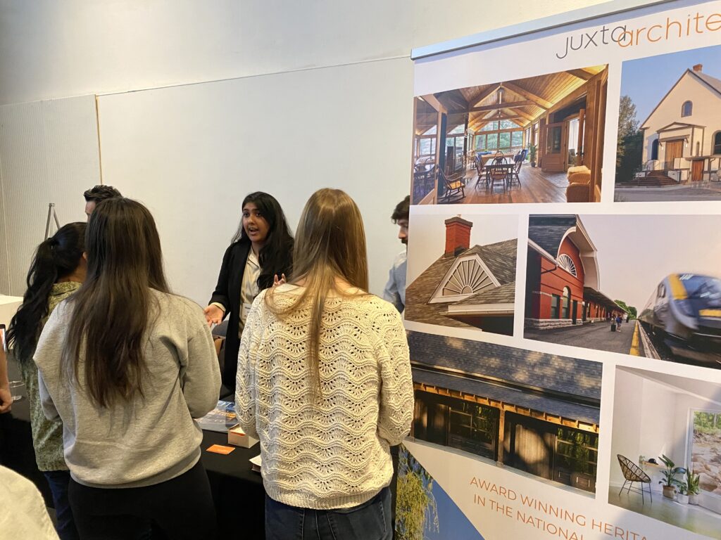 A representative from Juxta Architecture speaks with several students near a large project poster.