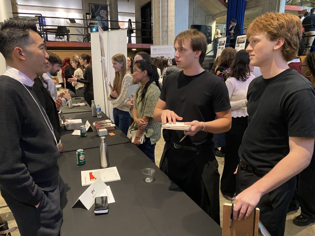 A firm representative talks with students at a table covered with booklets, water bottles, and name cards.