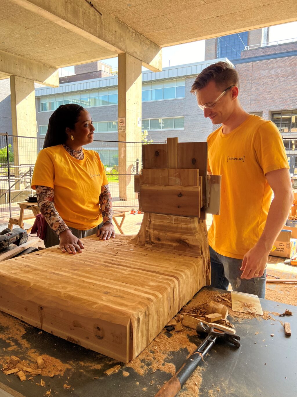 Two students standing at a workbench with a large wooden block between them in an outdoor covered area.