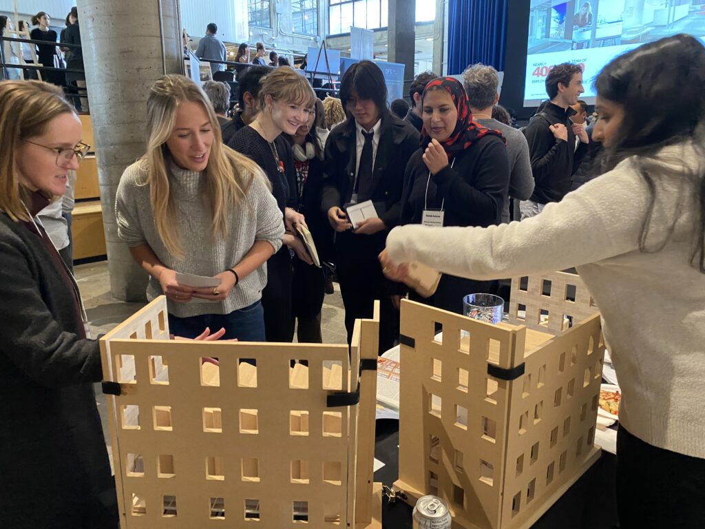 Students and firm representatives gather around a large wooden architectural model at a display table.