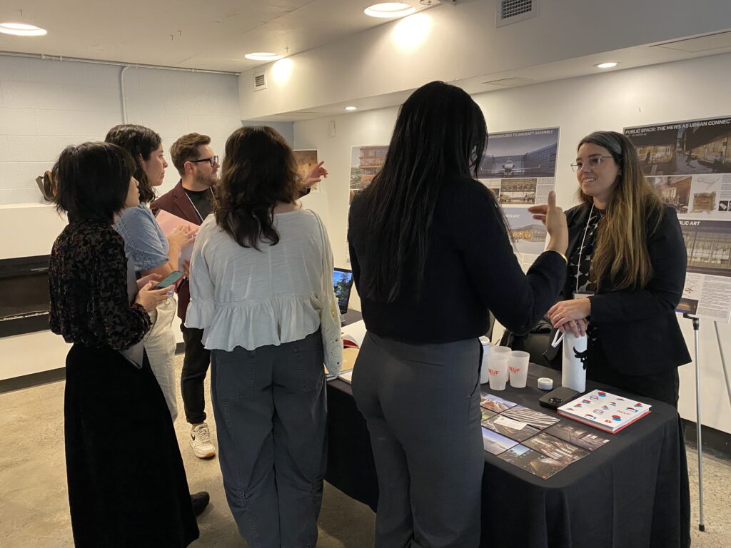 A group of students talk with a representative at a table displaying brochures and architectural project boards.
