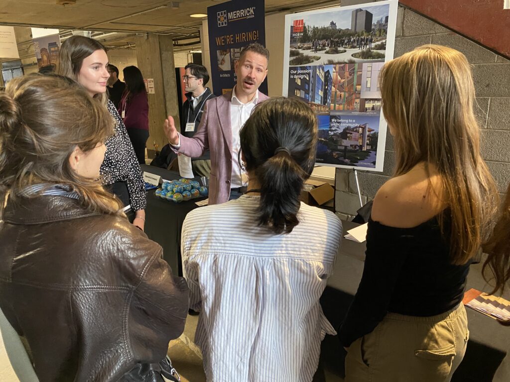 A firm representative speaks to a group of students in front of a Merrick poster and project boards.