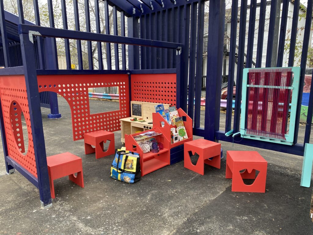 Red children’s play area with small stools, bookshelves, and a toy kitchen under a blue structure.