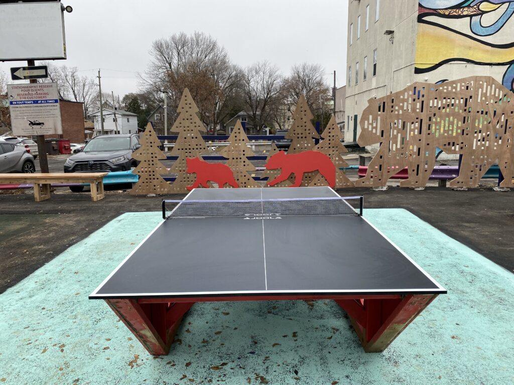 Outdoor ping pong table in front of cutout bear and tree silhouettes in the play area.