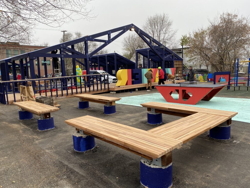 View of outdoor play and gathering structures with wooden benches, colorful panels, and people on site.
