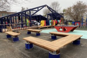 View of outdoor play and gathering structures with wooden benches, colorful panels, and people on site.