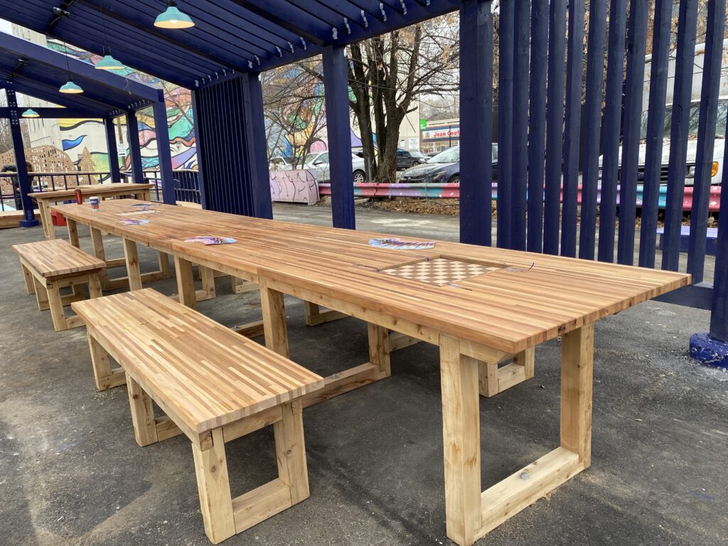 Long wooden tables and benches under a blue open-frame shelter with hanging lights.