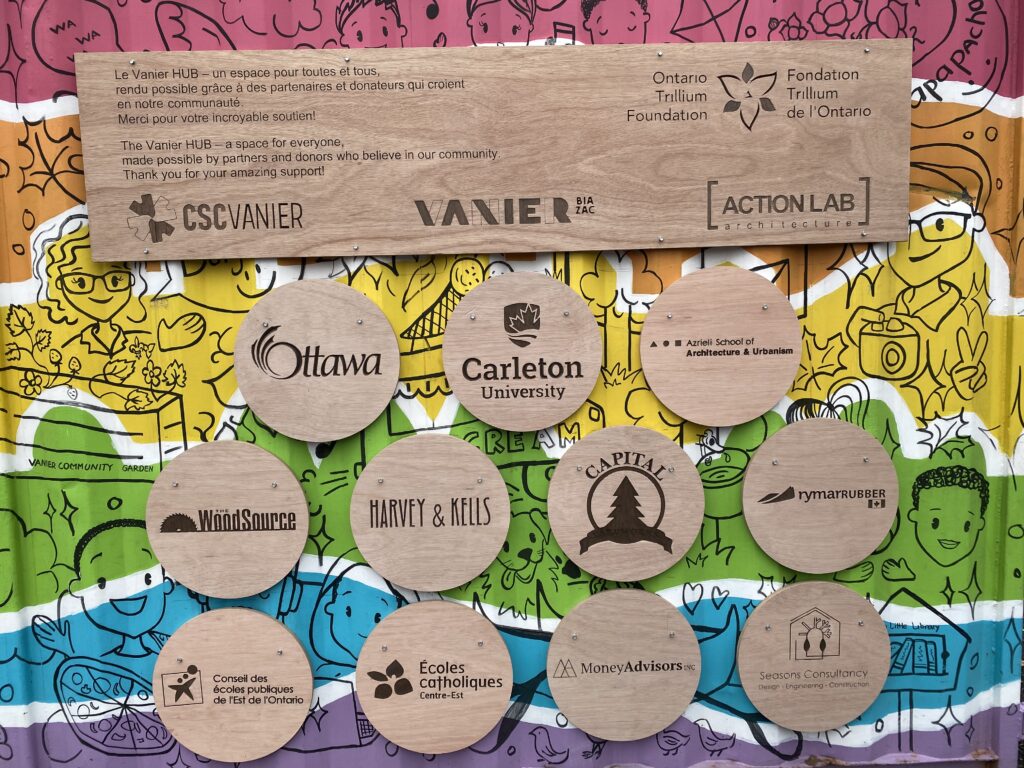 Wooden plaques with partner logos mounted on a colorful illustrated background.