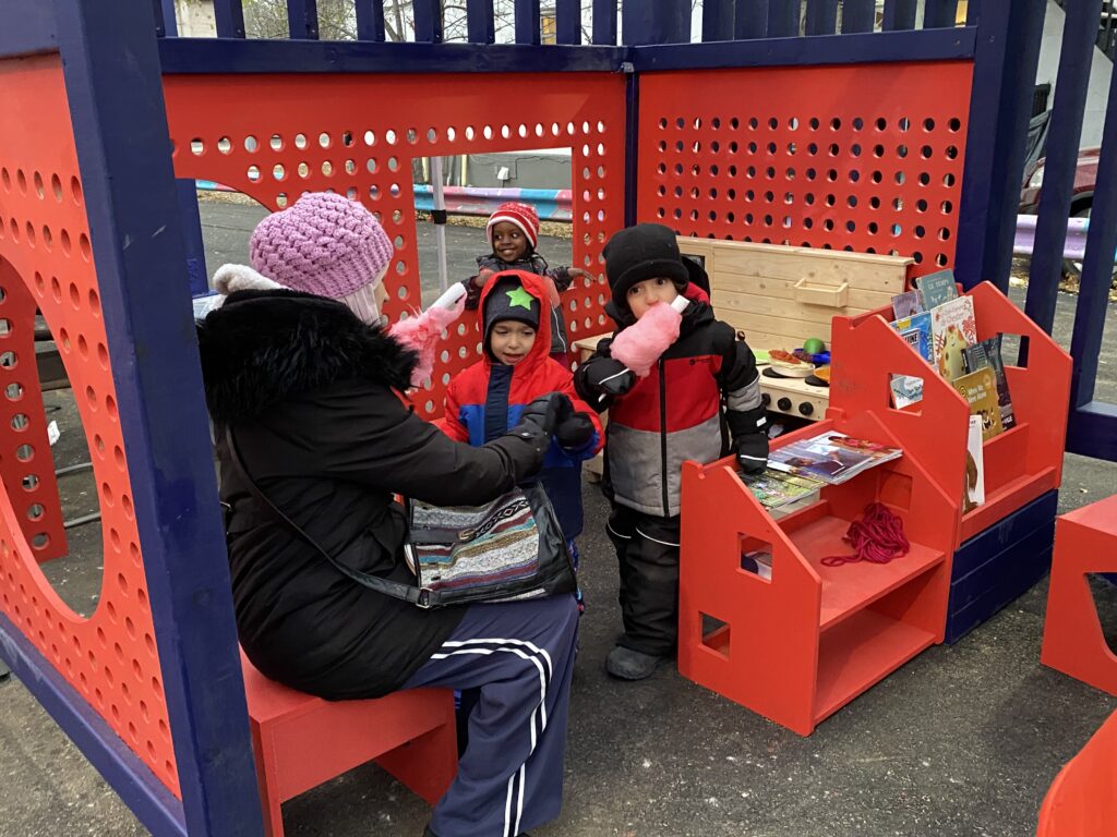 Children gathered in the red playhouse reading corner with small shelves, stools, and toys.