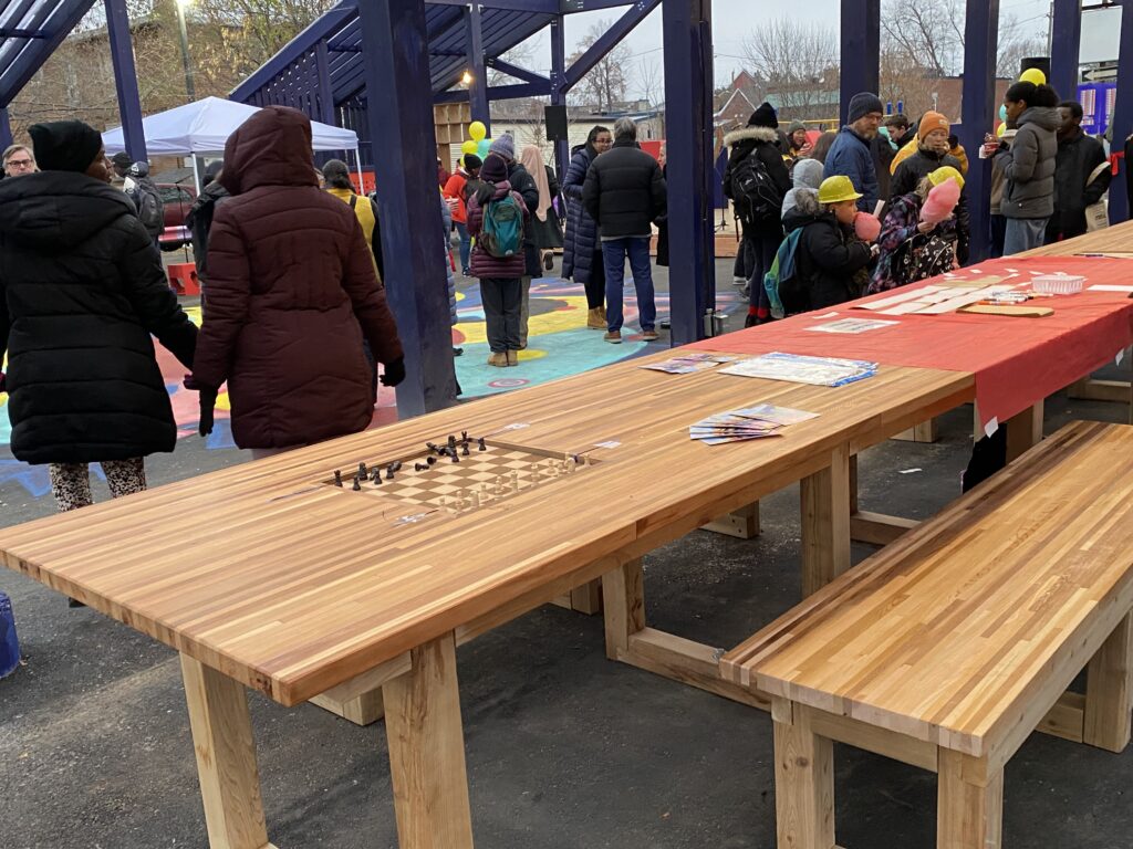 People gathering around the long wooden tables with a built-in chessboard under the blue-framed canopy.