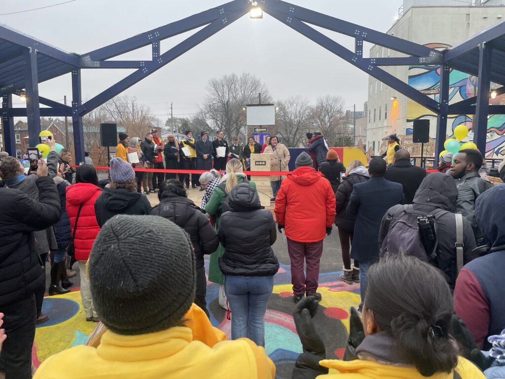 Community gathering at the Vanier Hub opening, with a speaker at the podium and a ribbon across the outdoor structure.