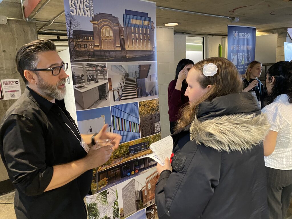 Student speaking with Laszlo Mohacsi, with a KWC Architects poster showing project images in the background.