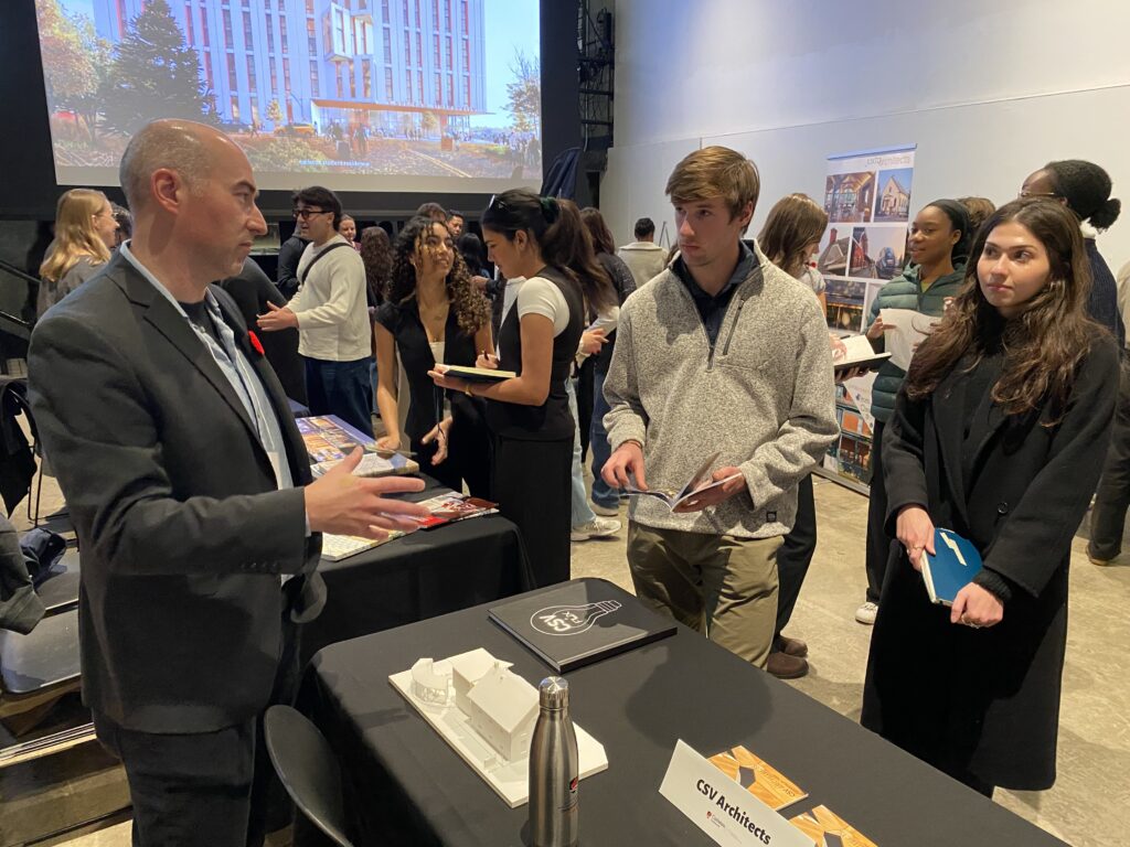 Rick Kellner speaking to two students in front of a display table with a white architectural model.