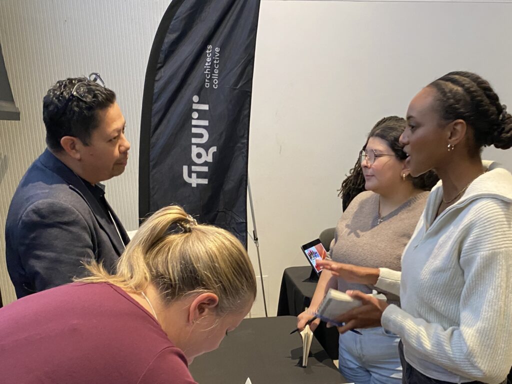 Three students speaking with Roberto Campos at the Figurr Architects Collective booth, with notebooks and phones in hand.