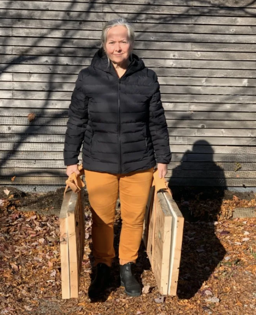 Sheryl Boyle standing outdoors holding two wooden wooden panels, with a wooden wall in the background.