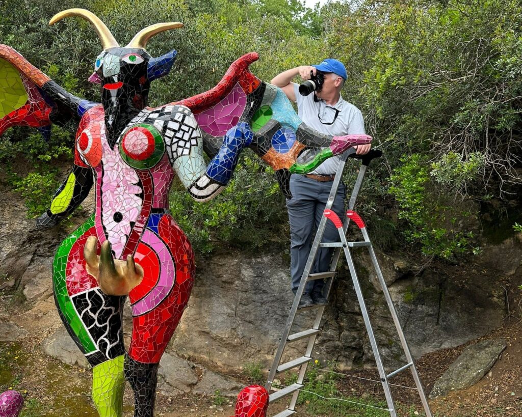Dr. Santana-Quintero standing on ladder photographing a large, colorful mosaic sculpture of a horned, winged figure.