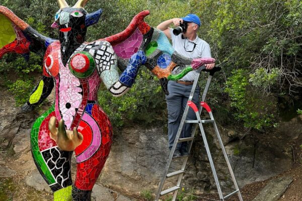 Dr. Santana-Quintero standing on ladder photographing a large, colorful mosaic sculpture of a horned, winged figure.