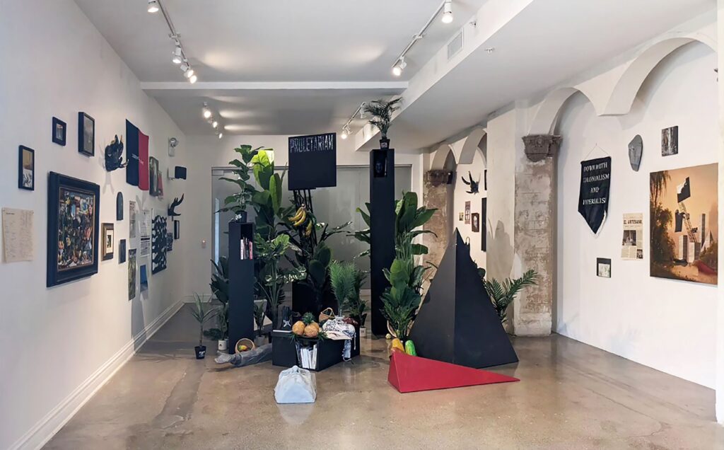 Gallery space filled with green plants and black sculptural forms, with wall-mounted text, images, and banners