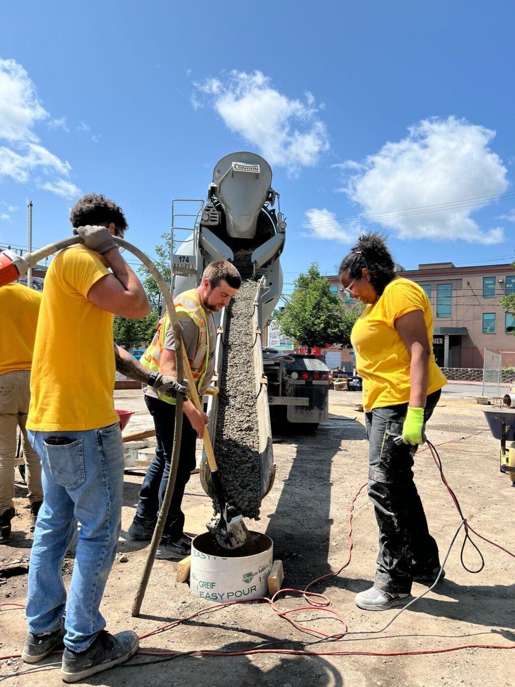 Menna Agha and students pouring concrete from a mixer into a cylindrical form on a sunny construction site.