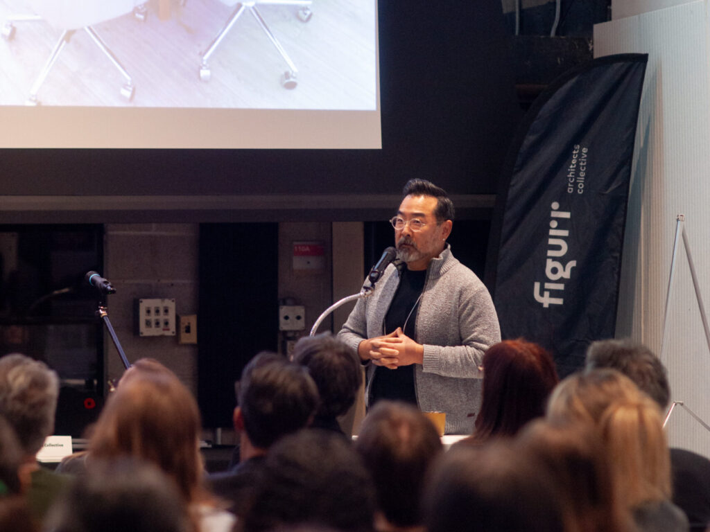 Raymon Yim giving a presentation on stage in front of a large screen showing a photo of a design meeting.