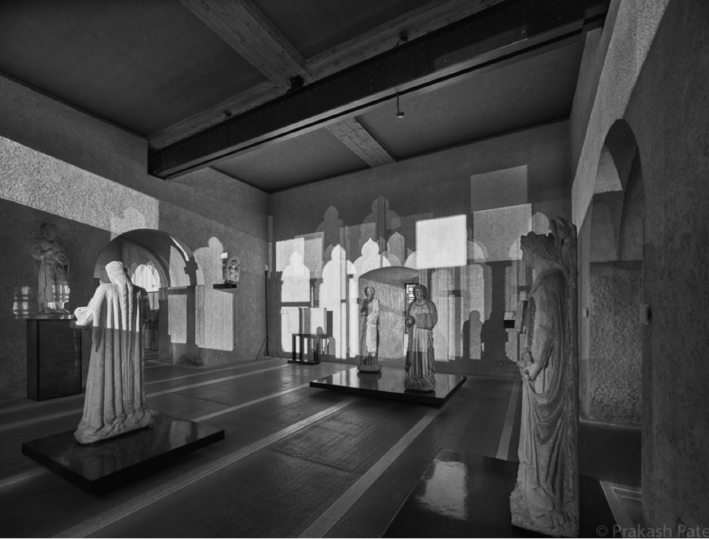 Museum gallery with sculptural figures on plinths, arranged within a concrete interior.