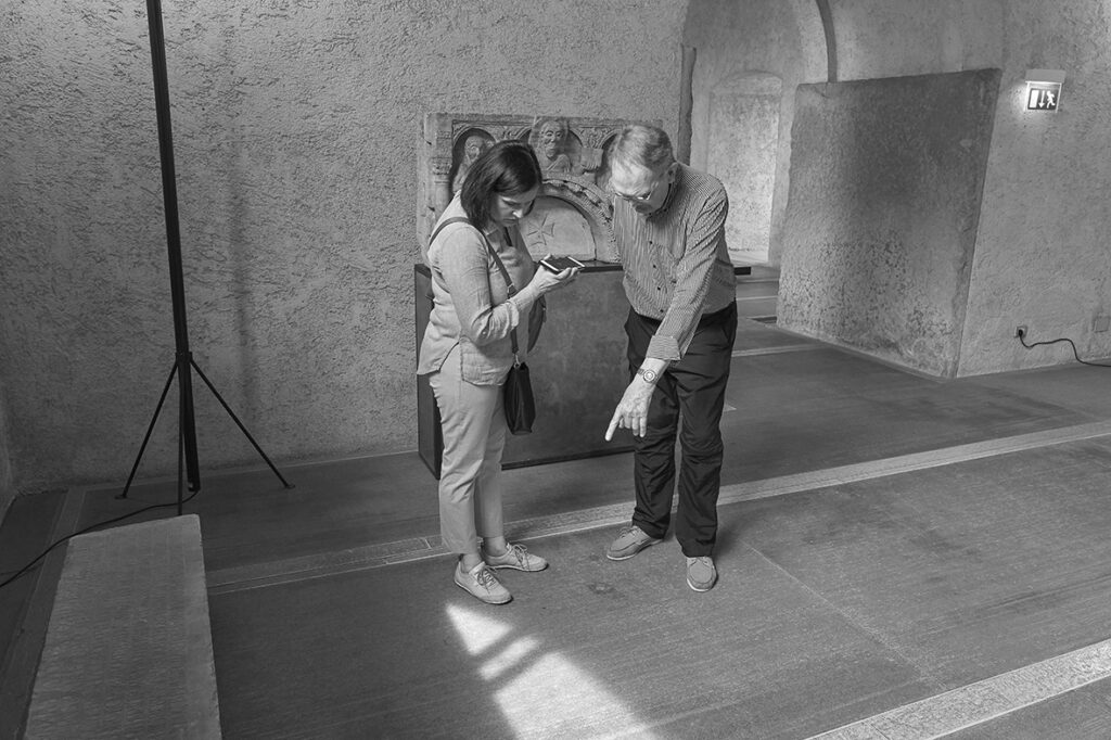 Two people standing indoors, looking down at a narrow strip of light on a stone floor.