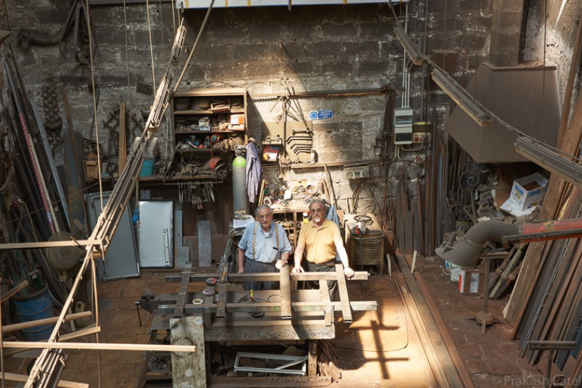 Two people standing at a workbench inside a workshop filled with tools, materials, and machinery.